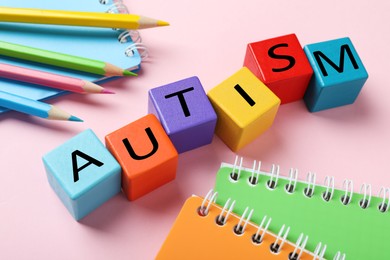 Photo of Word Autism made with colorful cubes, pencils and notebooks on pink background, closeup