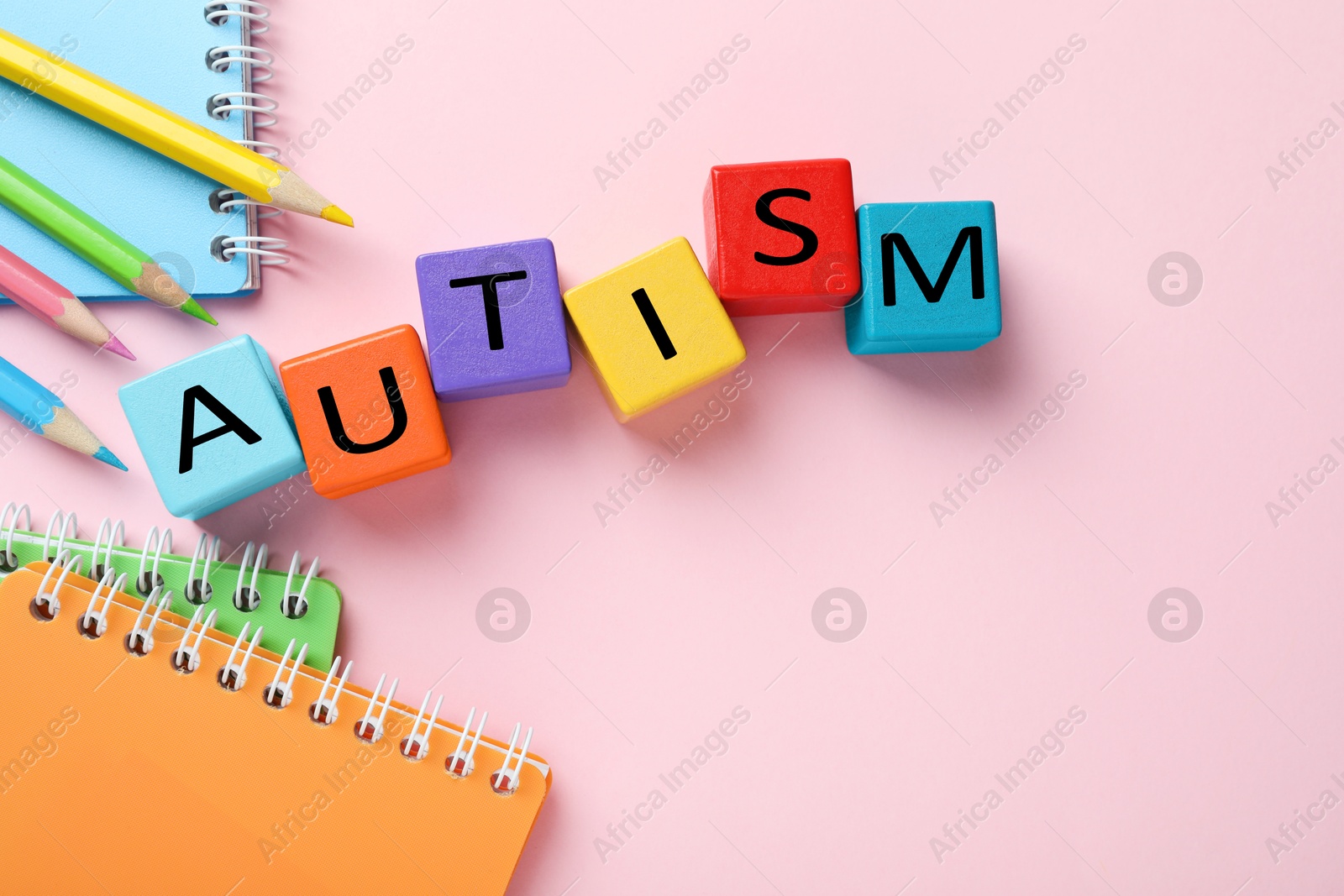 Photo of Word Autism made with colorful cubes, pencils and notebooks on pink background, flat lay