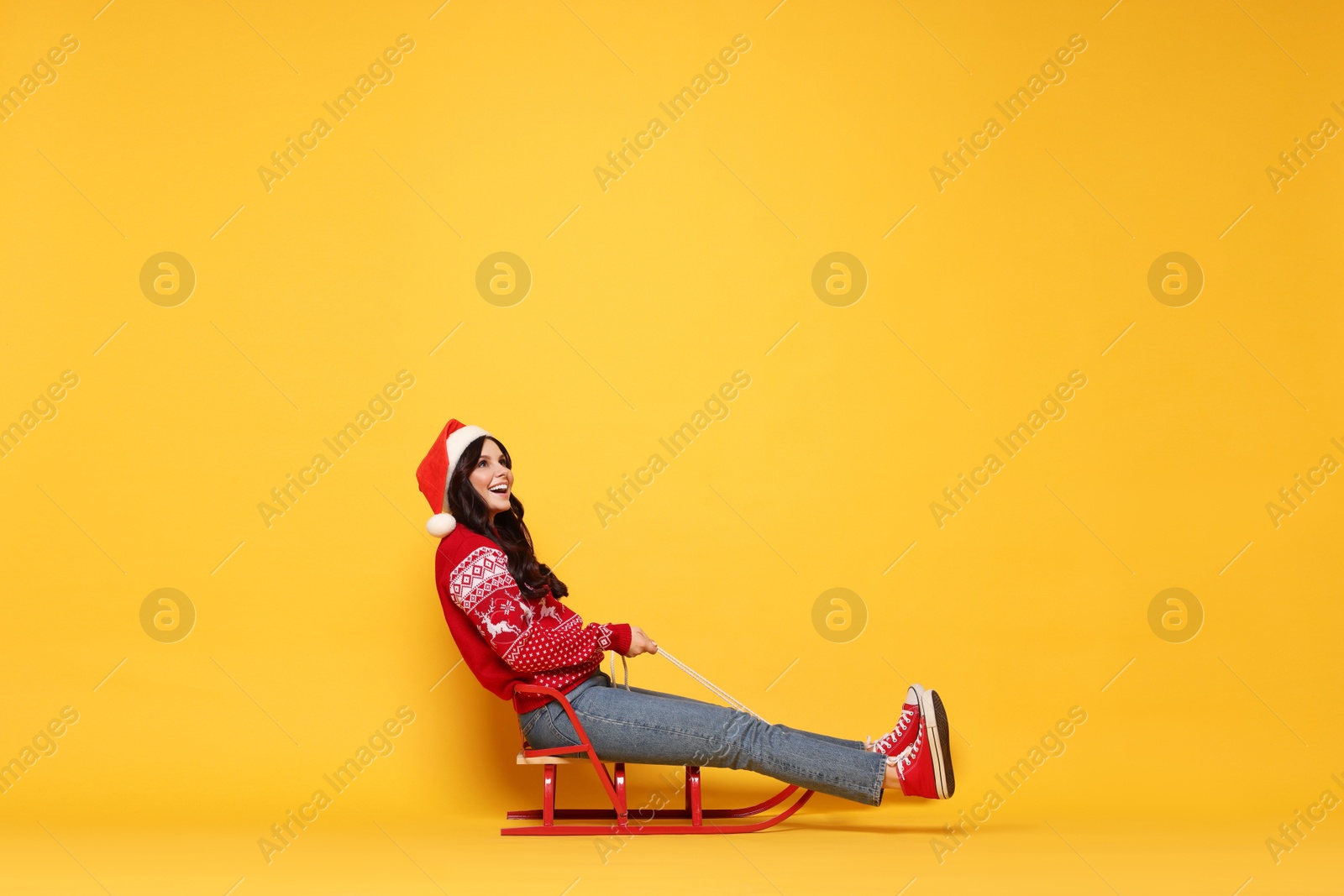 Photo of Woman in Santa hat on sledge against yellow background, space for text. Christmas celebration