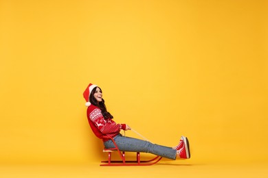 Woman in Santa hat on sledge against yellow background, space for text. Christmas celebration