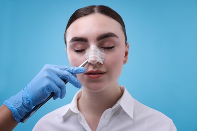 Photo of Doctor removing medical bandage from patient's nose after plastic surgery operation on light blue background, closeup