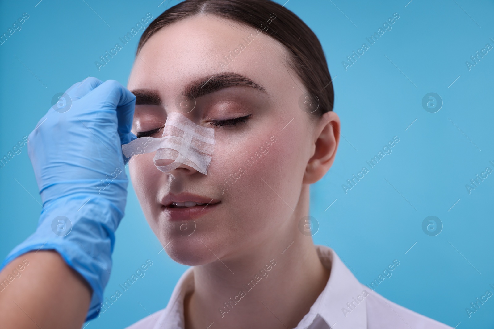 Photo of Doctor removing medical bandage from patient's nose after plastic surgery operation on light blue background, closeup