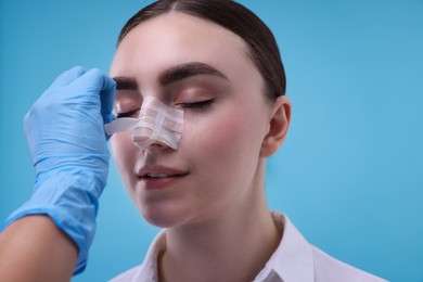 Photo of Doctor removing medical bandage from patient's nose after plastic surgery operation on light blue background, closeup