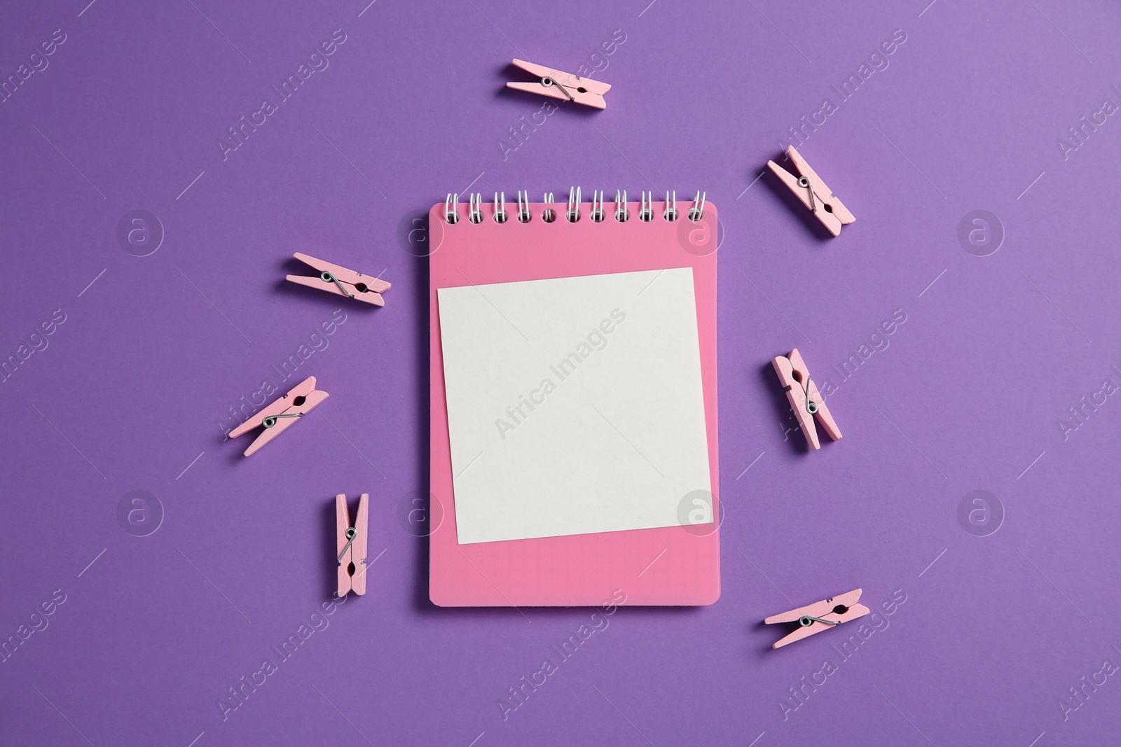 Photo of Blank note, notebook and clothing pegs on purple background, flat lay. Space for text