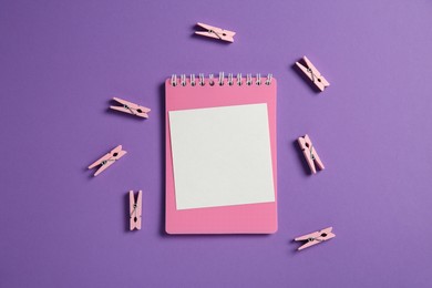 Photo of Blank note, notebook and clothing pegs on purple background, flat lay. Space for text