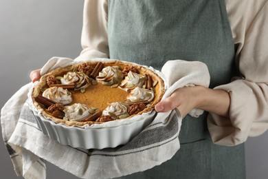 Photo of Woman holding homemade pumpkin pie with whipped cream, seeds and cinnamon on grey background, closeup