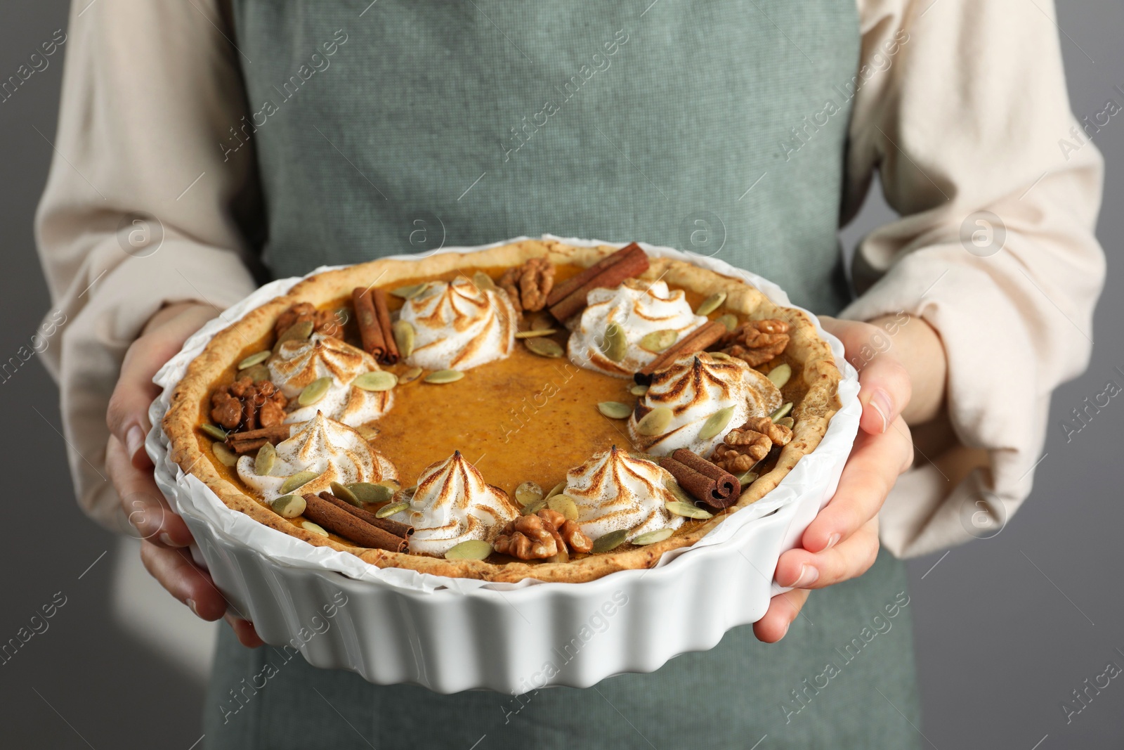 Photo of Woman holding homemade pumpkin pie with whipped cream, seeds and cinnamon on grey background, closeup