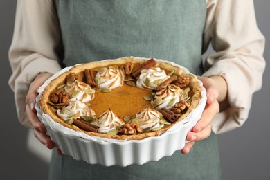 Photo of Woman holding homemade pumpkin pie with whipped cream, seeds and cinnamon on grey background, closeup