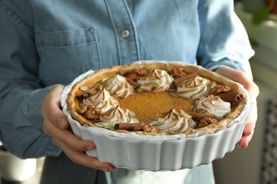 Photo of Woman holding homemade pumpkin pie with whipped cream, seeds and cinnamon on blurred background, closeup