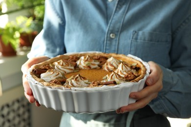 Photo of Woman holding homemade pumpkin pie with whipped cream, seeds and cinnamon on blurred background, closeup