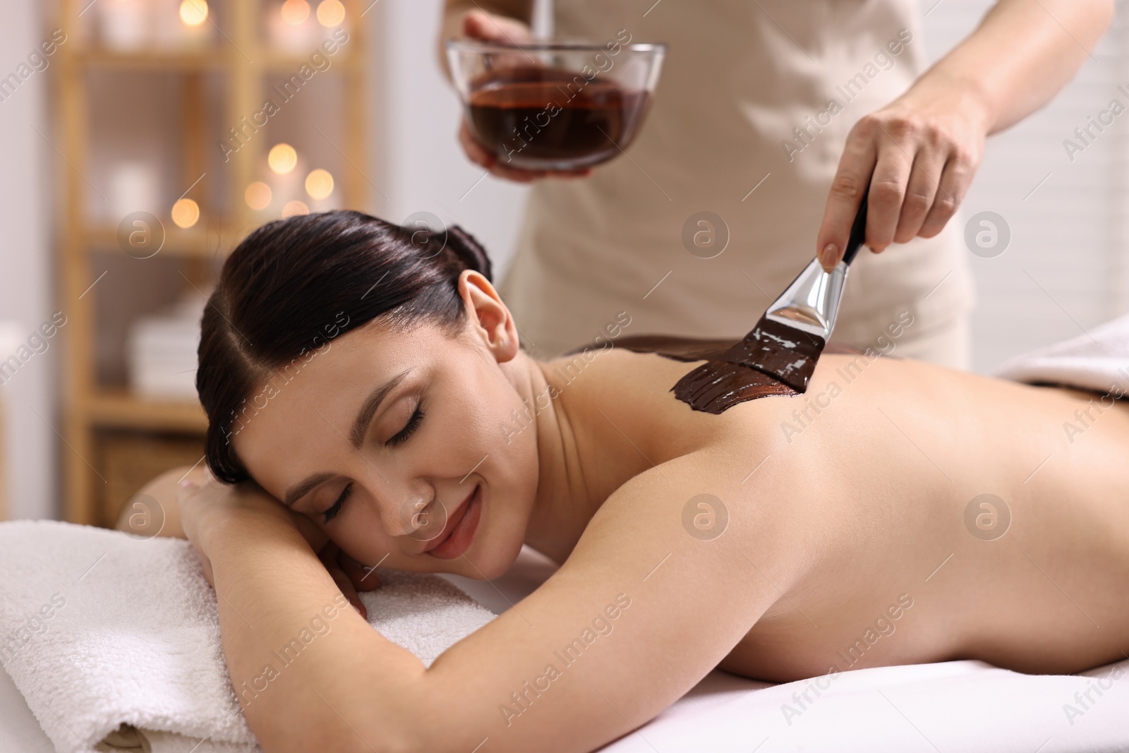 Photo of Chocolate body wrap. Spa worker applying mask onto woman's back in salon, closeup