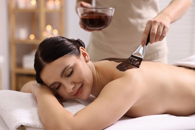 Photo of Chocolate body wrap. Spa worker applying mask onto woman's back in salon, closeup