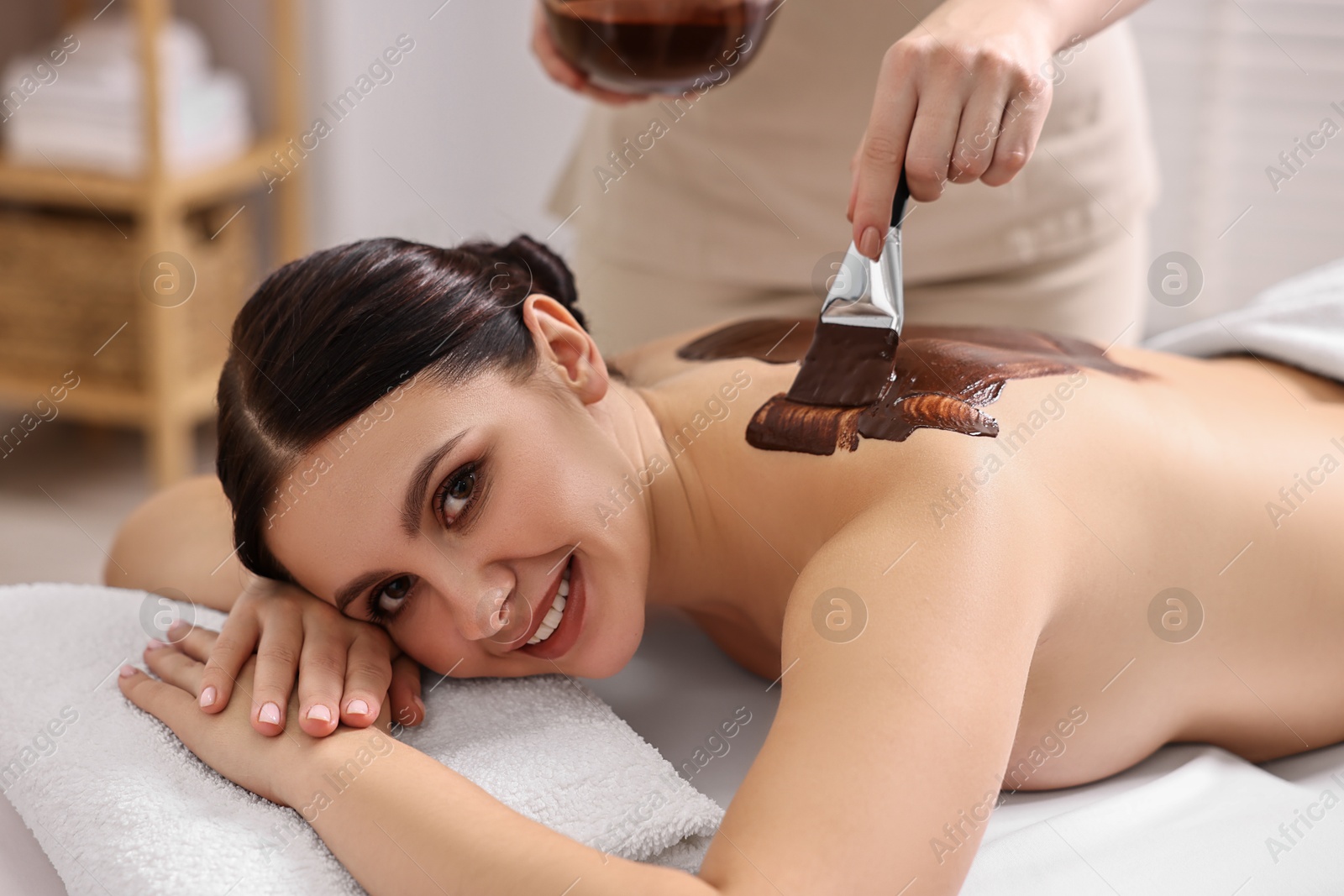 Photo of Chocolate body wrap. Spa worker applying mask onto woman's back in salon, closeup