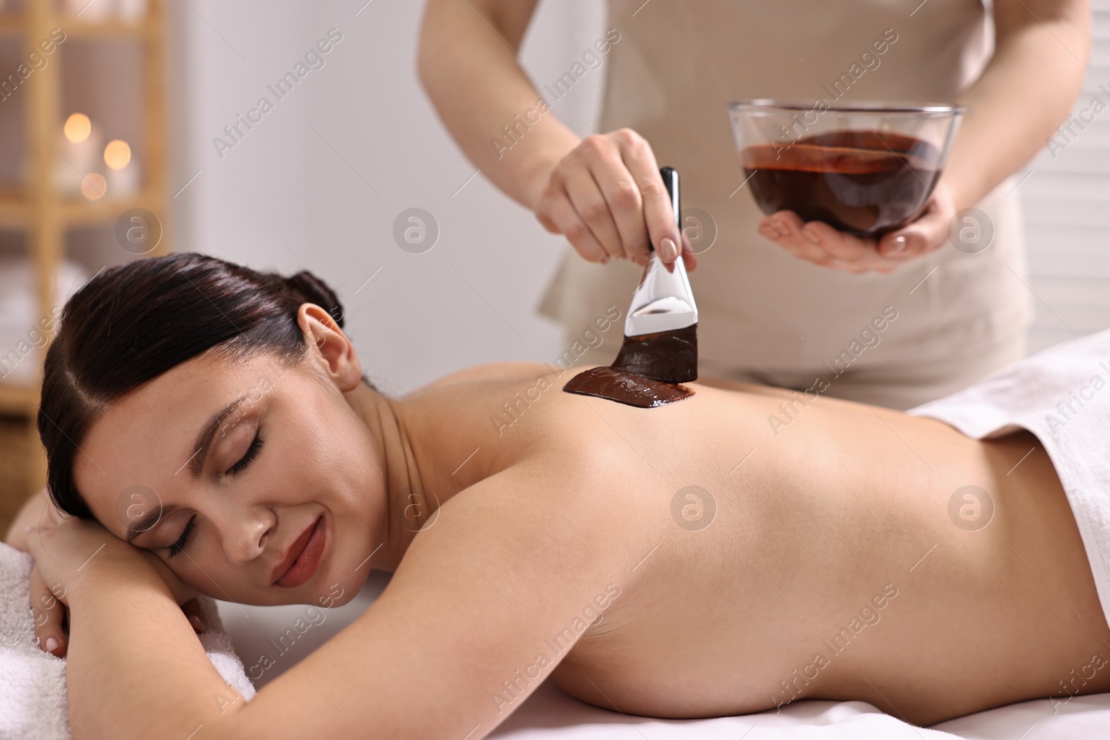 Photo of Chocolate body wrap. Spa worker applying mask onto woman's back in salon, closeup