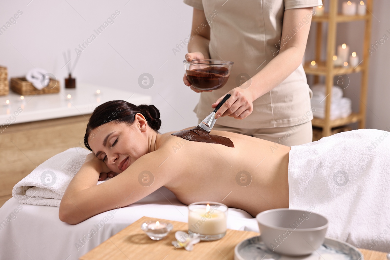 Photo of Chocolate body wrap. Spa worker applying mask onto woman's back in salon, closeup