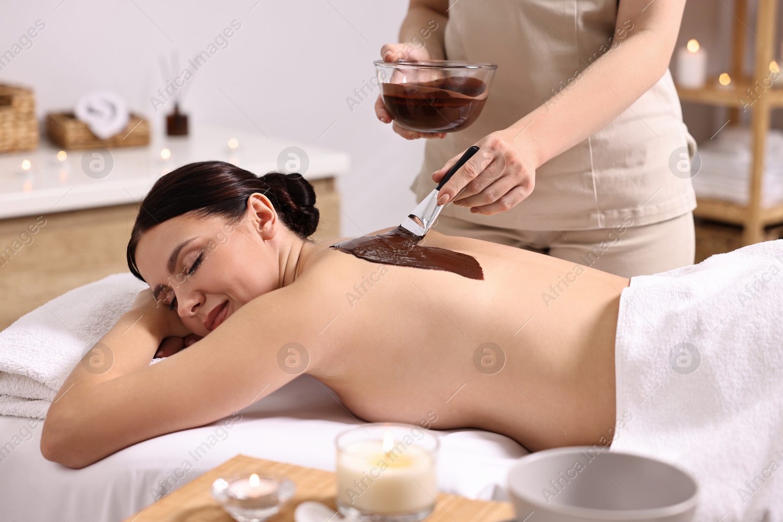 Photo of Chocolate body wrap. Spa worker applying mask onto woman's back in salon, closeup