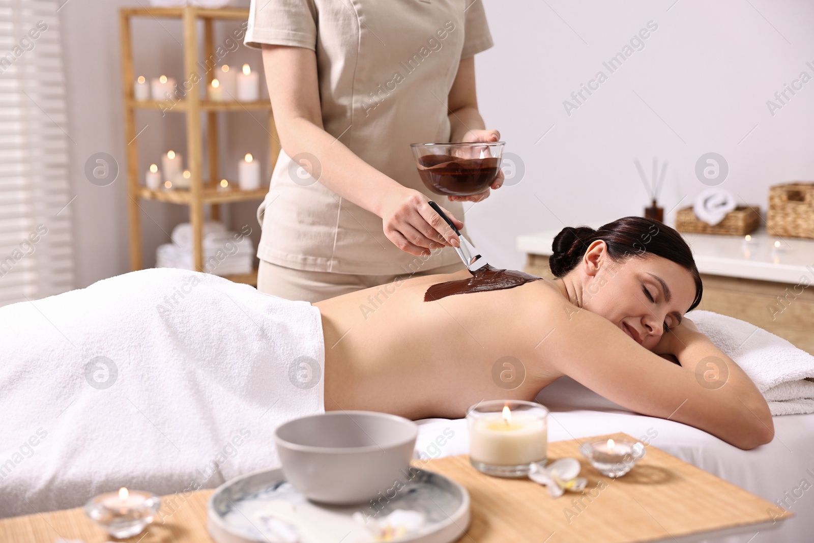 Photo of Chocolate body wrap. Spa worker applying mask onto woman's back in salon, closeup