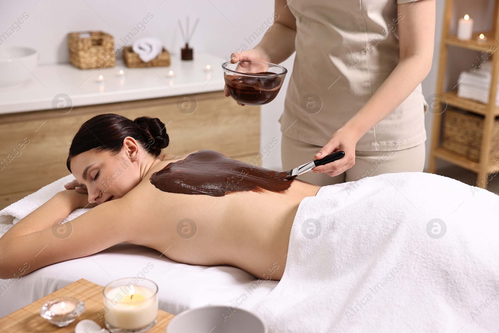 Photo of Chocolate body wrap. Spa worker applying mask onto woman's back in salon, closeup