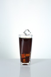 Photo of Ice cube falling into glass with tasty cola on mirror table against white background