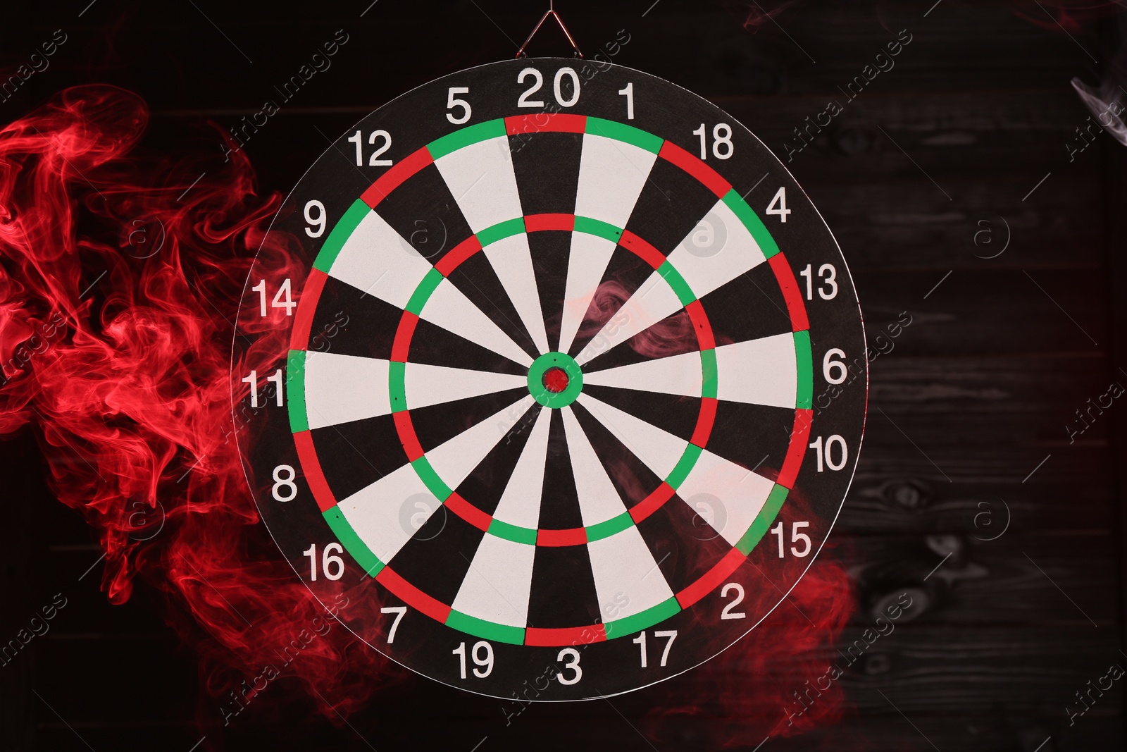 Photo of Dart board hanging against black wooden background in color light and smoke