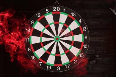 Photo of Dart board hanging against black wooden background in color light and smoke