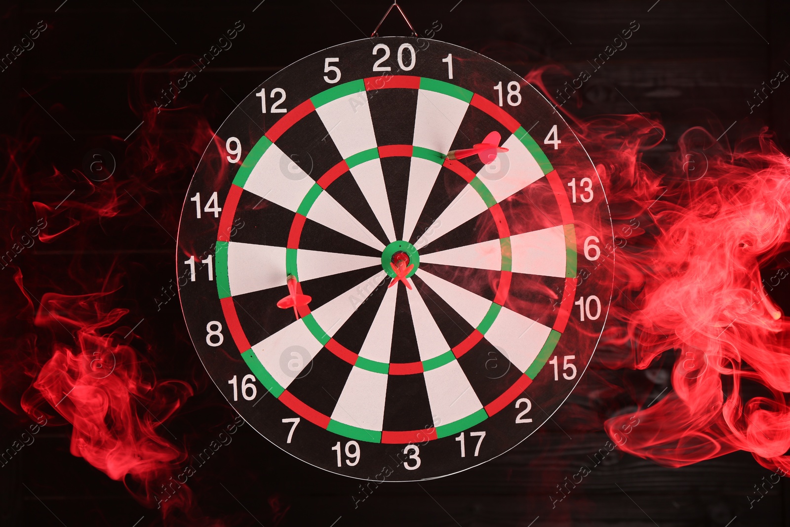 Photo of Dart board with red arrow hanging against black wooden background in color light and smoke