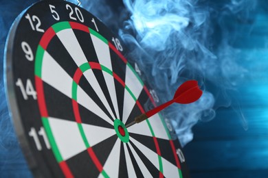 Photo of Dart board with red arrow against blurred blue background in smoke, closeup