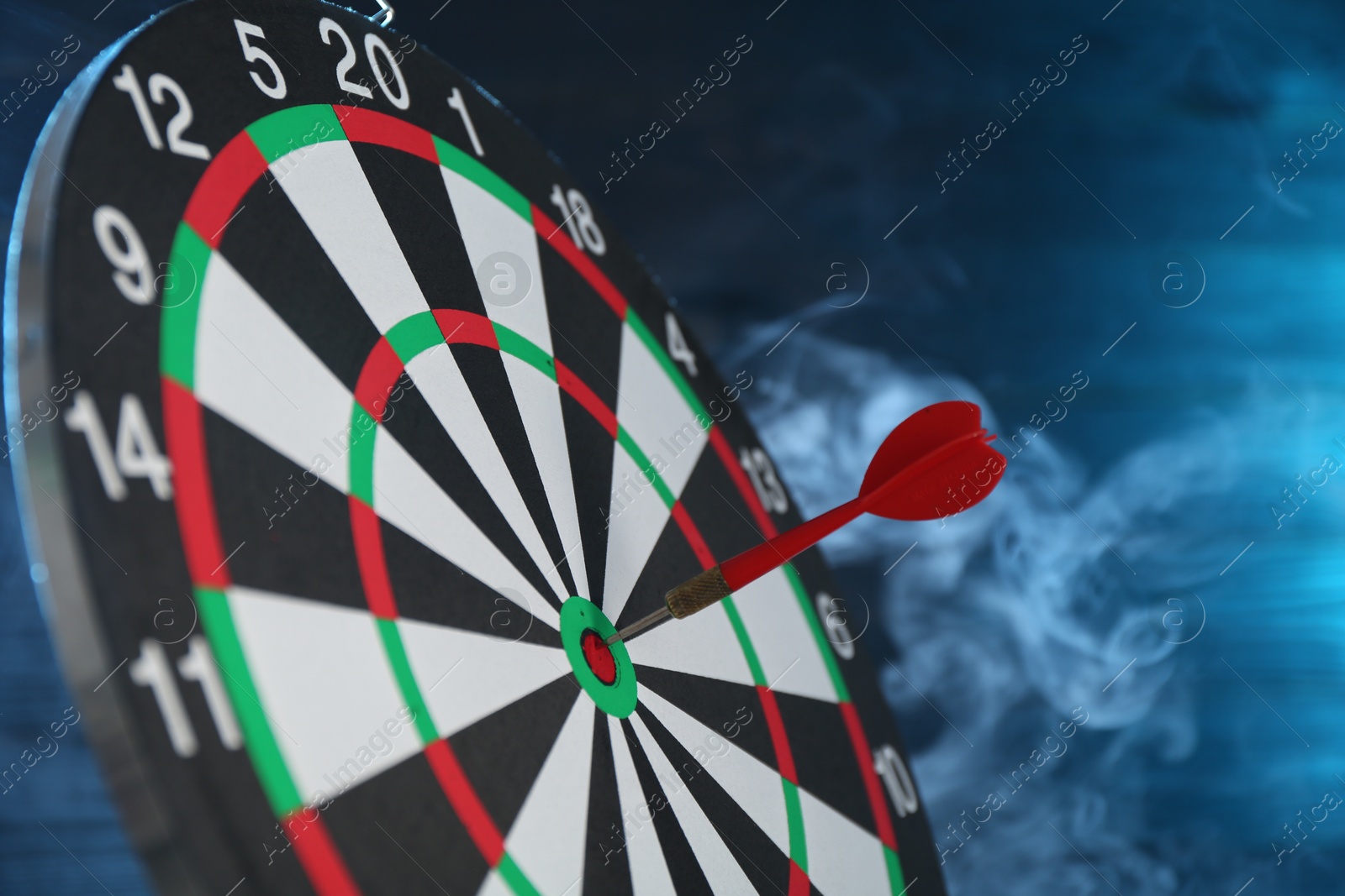 Photo of Dart board with red arrow against blurred blue background in smoke, closeup