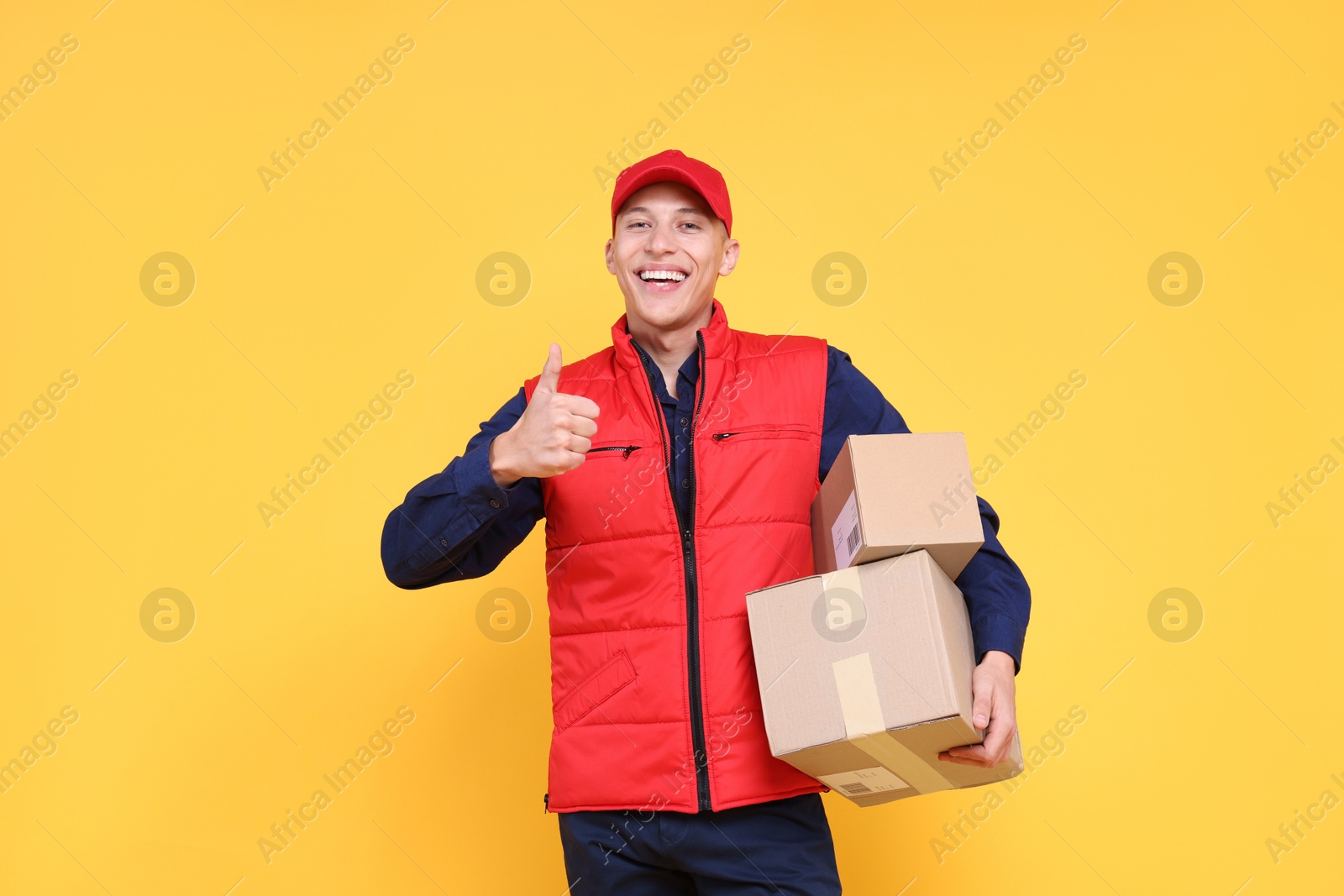 Photo of Happy postman with parcels showing thumbs up on yellow background