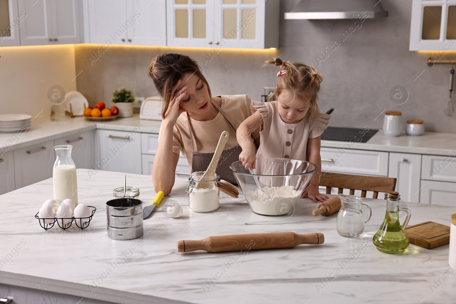 Photo of Tired housewife cooking with her little daughter at marble table in kitchen