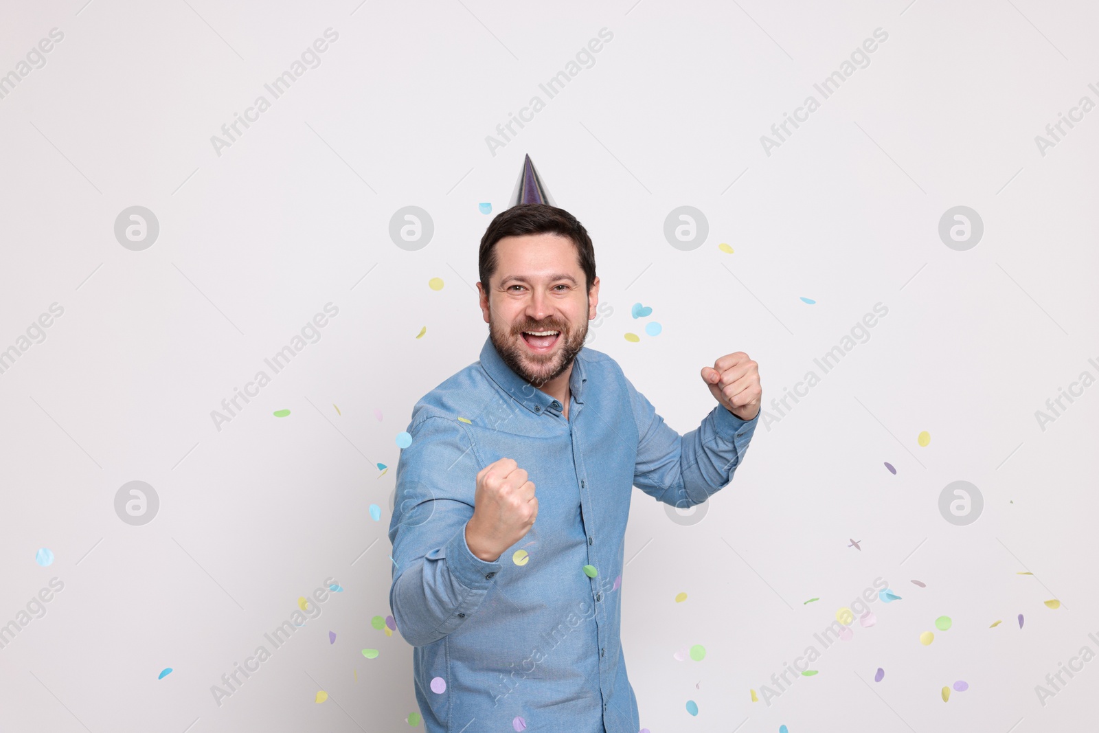 Photo of Happy man in conical paper hat and falling confetti on white background. Surprise party