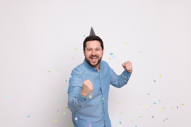 Happy man in conical paper hat and falling confetti on white background. Surprise party