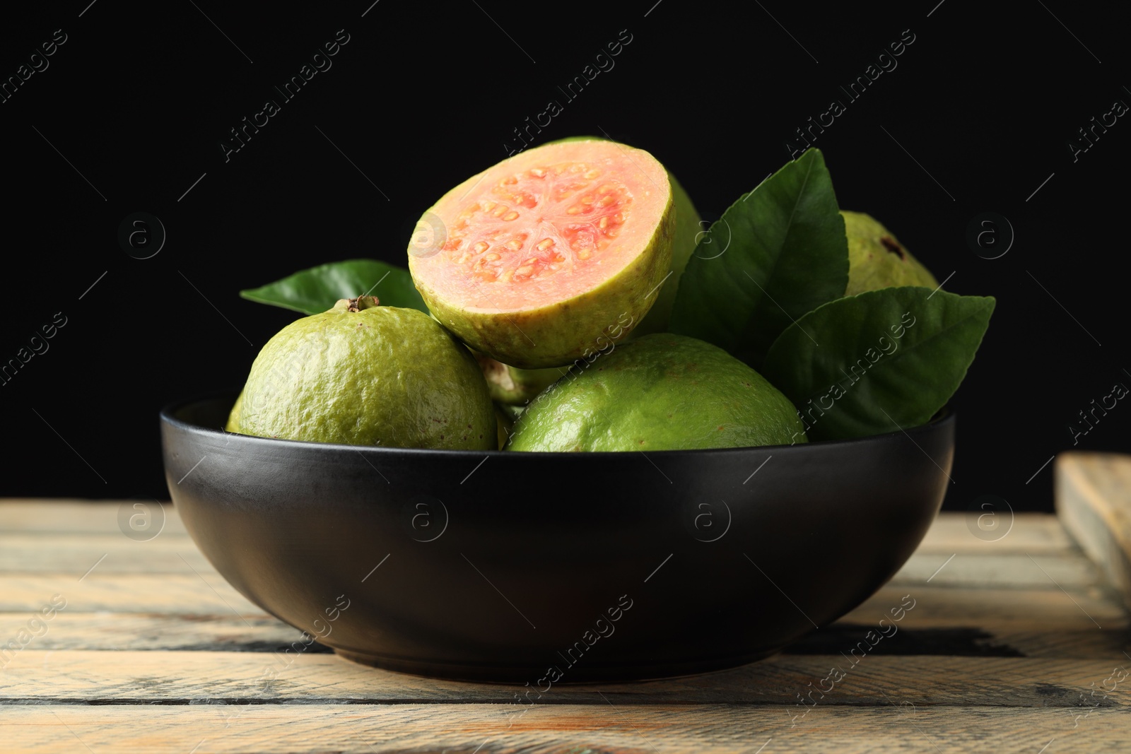 Photo of Fresh guava fruits in bowl on wooden table against black background, closeup