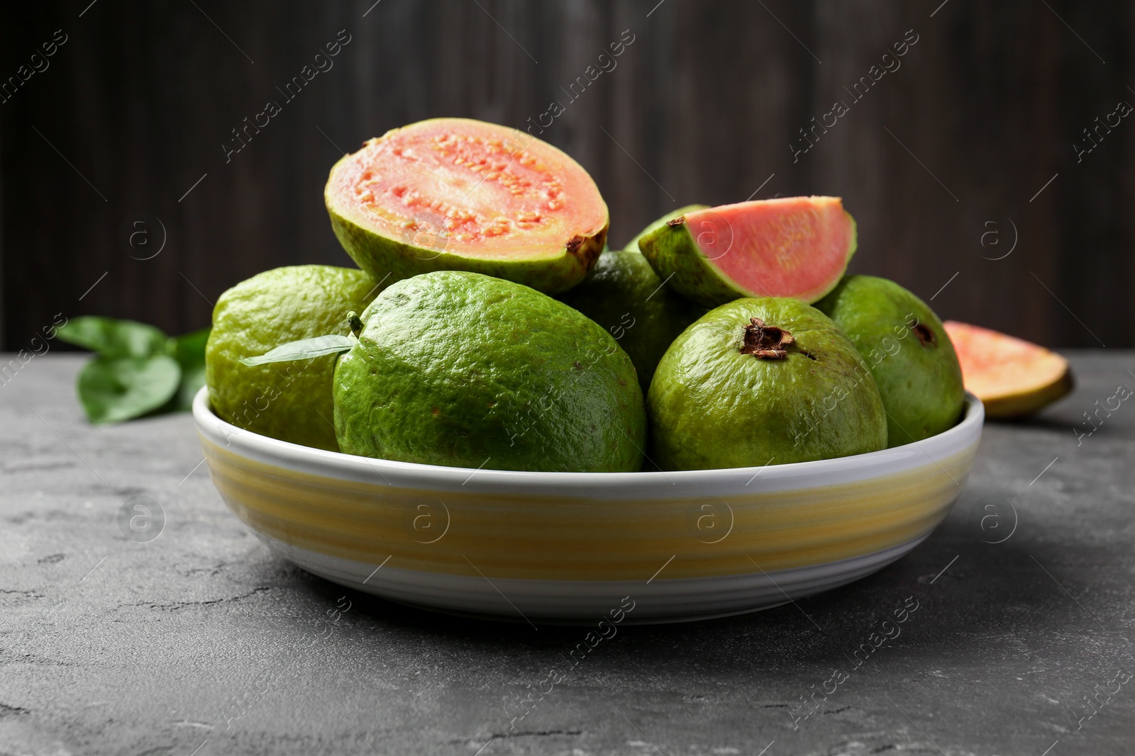 Photo of Fresh whole and cut guava fruits in bowl on grey textured table, closeup