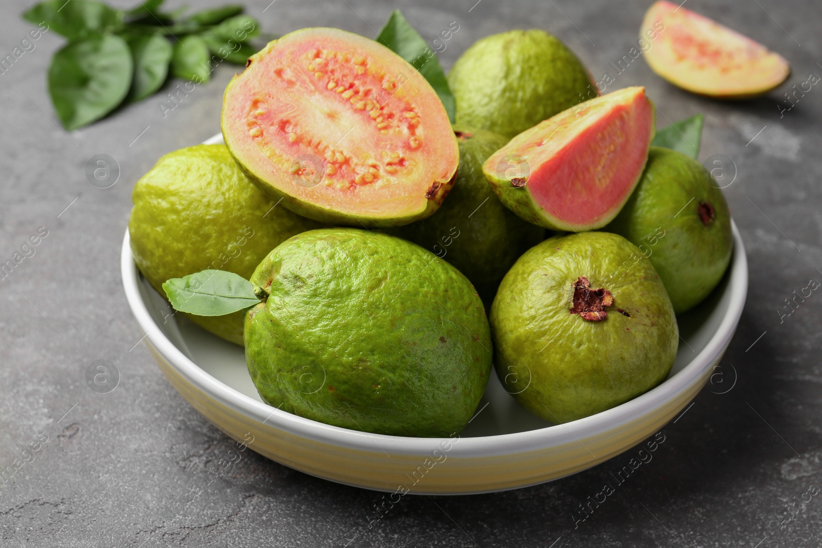 Photo of Fresh whole and cut guava fruits in bowl on grey textured table, closeup