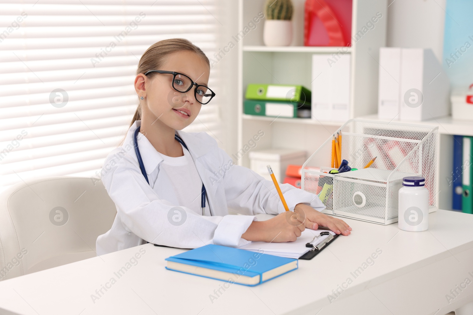 Photo of Little girl with stethoscope and clipboard pretending to be doctor at white table indoors