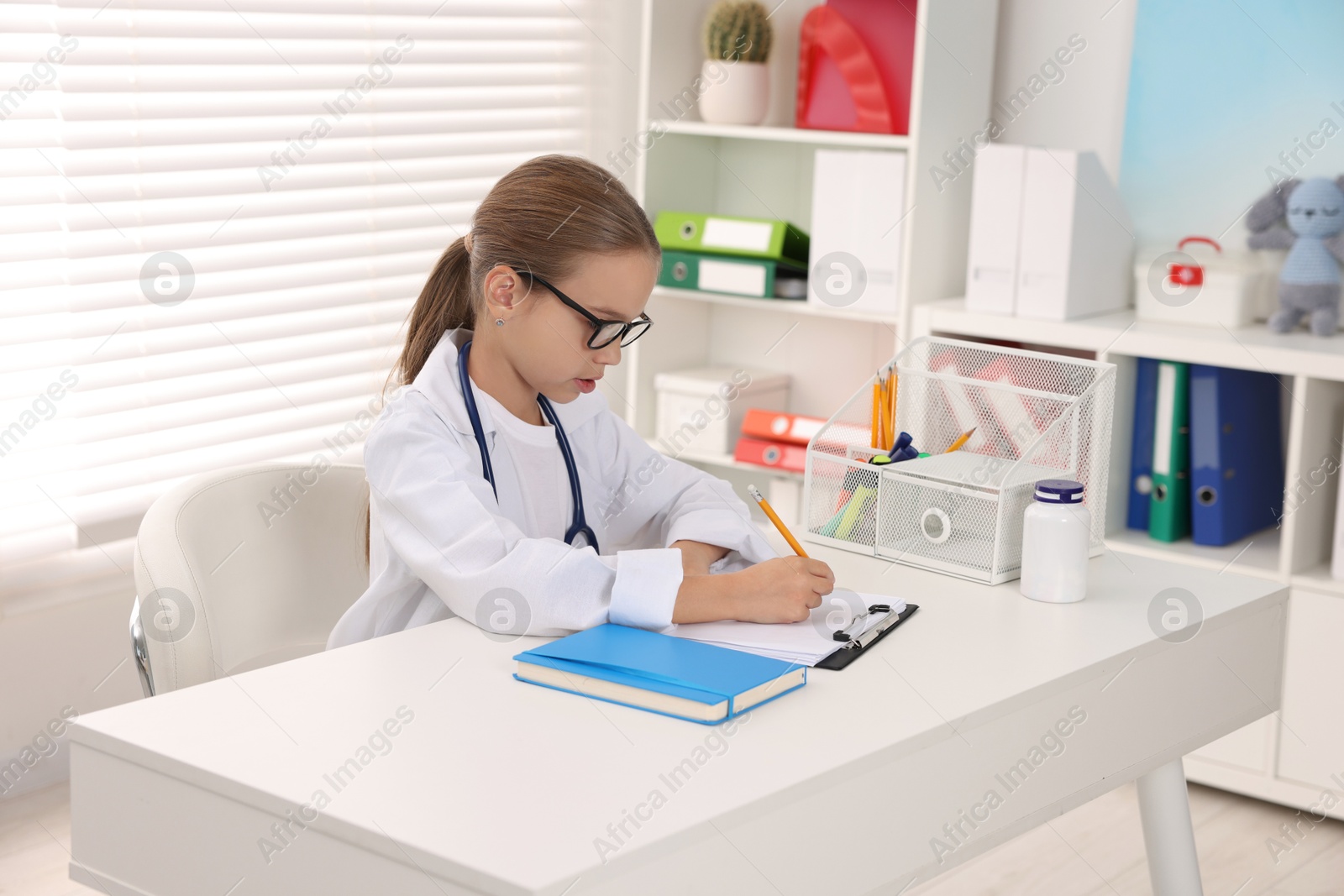 Photo of Little girl with stethoscope and clipboard pretending to be doctor at white table indoors