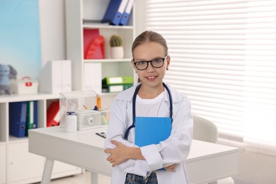 Photo of Girl with stethoscope and book pretending to be doctor indoors