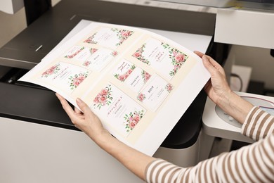 Photo of Woman with printed invitation cards near modern printer indoors, closeup. Printing house