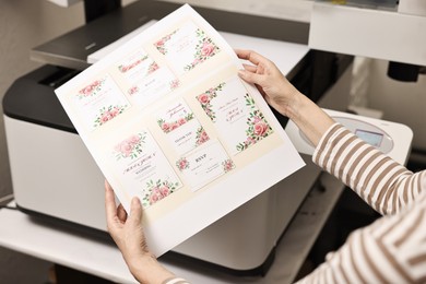 Photo of Woman with printed invitation cards near modern printer indoors, closeup. Printing house