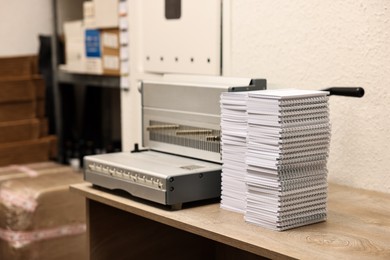 Photo of Modern binding machine and stacks of notebooks on wooden table indoors