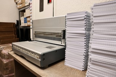 Photo of Modern binding machine and stacks of notebooks on wooden table indoors