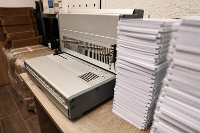 Photo of Modern binding machine and stacks of notebooks on wooden table indoors