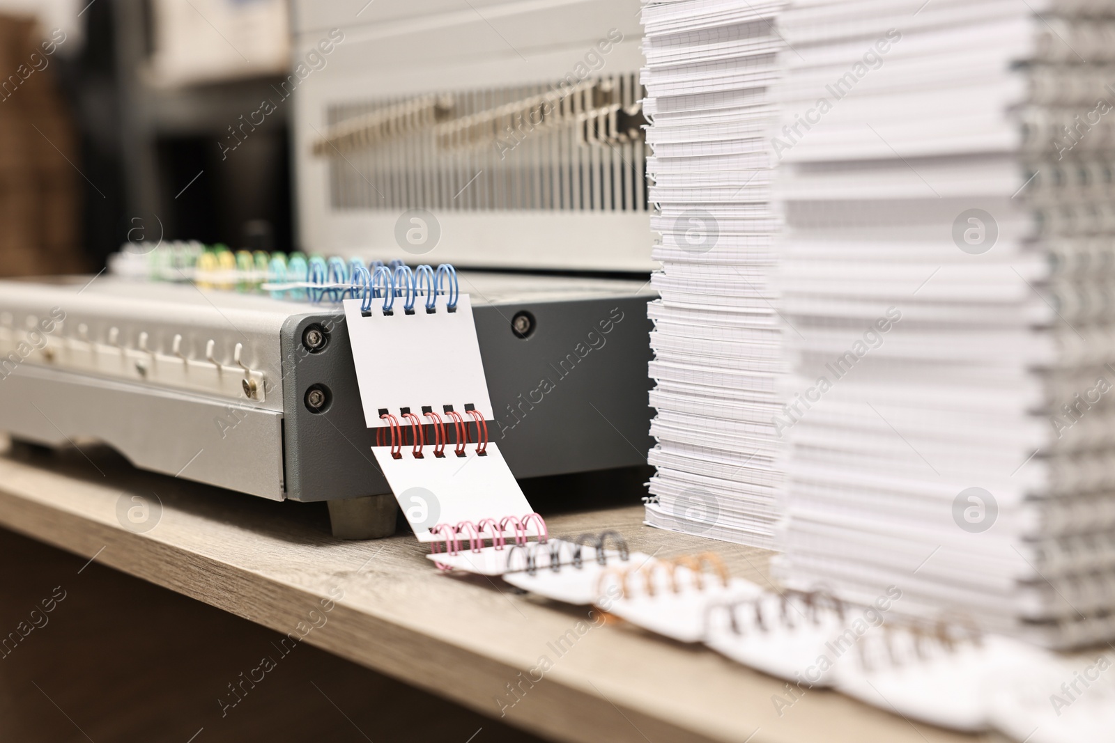 Photo of Modern binding machine, stacks of notebooks and double loop wire binding spines on wooden table indoors, closeup