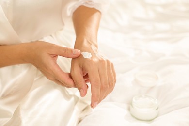 Photo of Woman applying cream onto hand on bed at home, closeup