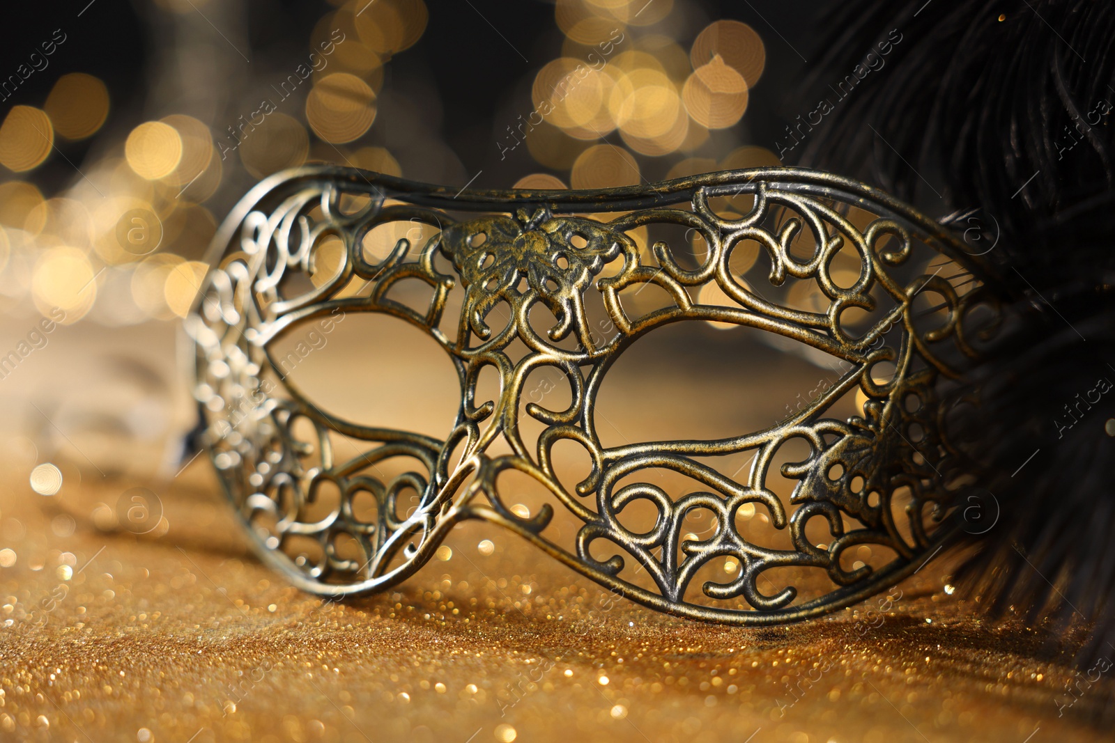 Photo of Beautiful carnival mask and black feathers on golden background against blurred lights, closeup. Bokeh effect