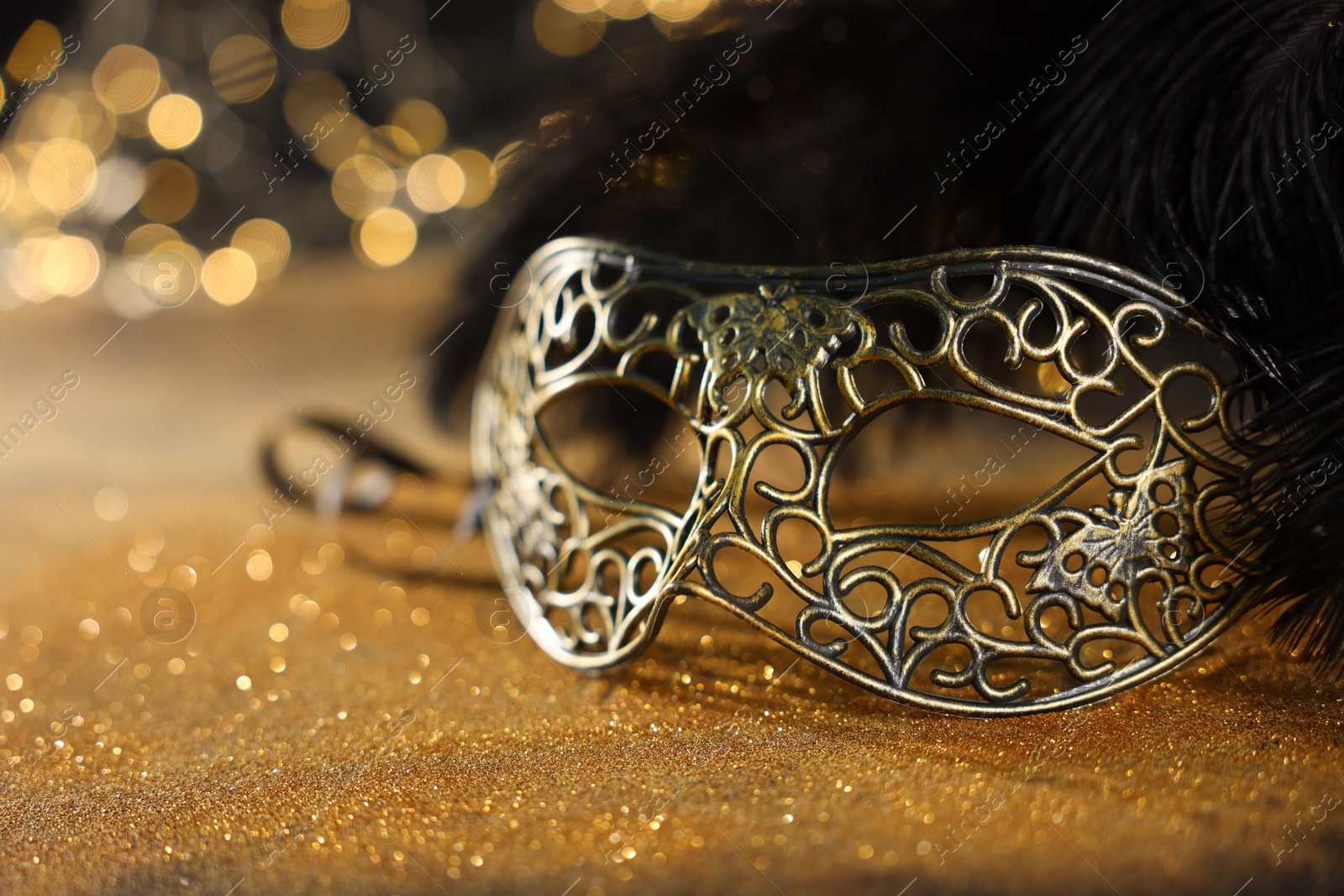 Photo of Beautiful carnival mask and black feathers on golden background against blurred lights, closeup. Bokeh effect