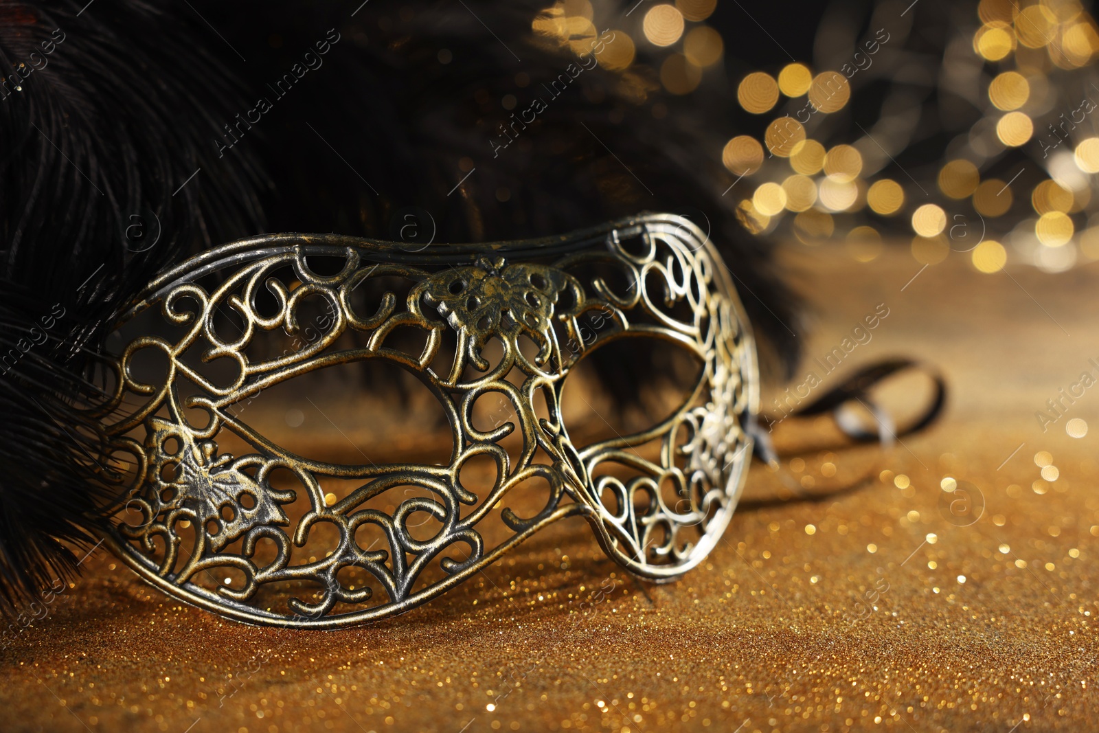 Photo of Beautiful carnival mask and black feathers on golden background against blurred lights, closeup. Bokeh effect