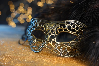 Photo of Beautiful carnival mask and black feathers on golden background against blurred lights, closeup. Bokeh effect