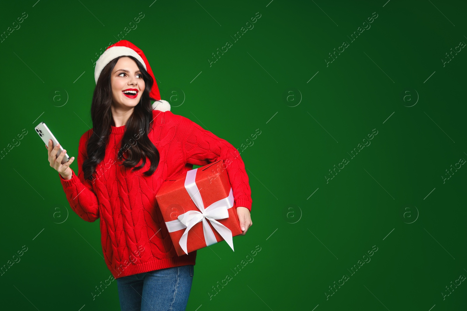 Photo of Attractive woman in Santa hat with Christmas gift and smartphone on green background, space for text